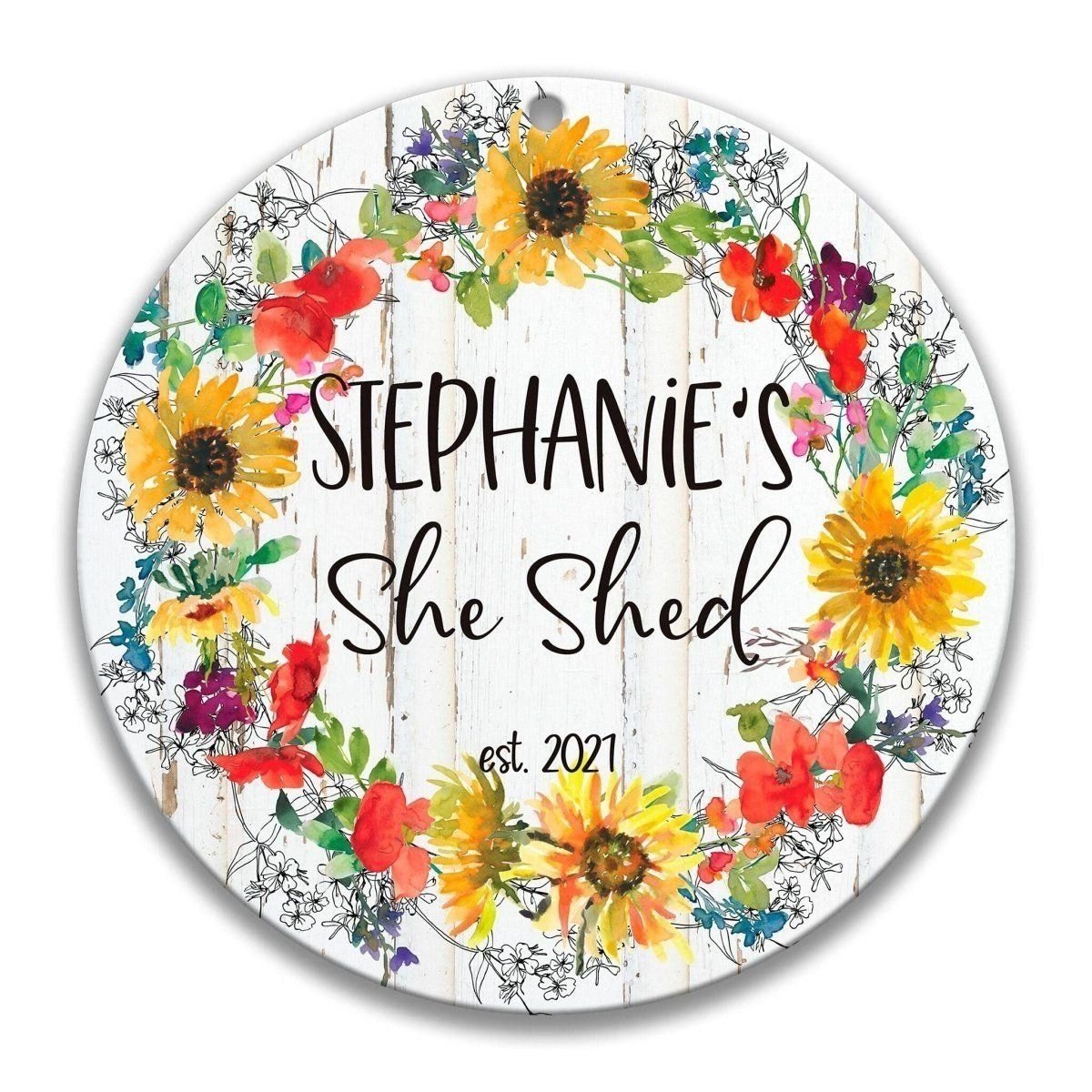 A round, white sign with a wreath of colorful flowers surrounding the text "Stephanie's She Shed est. 2021".