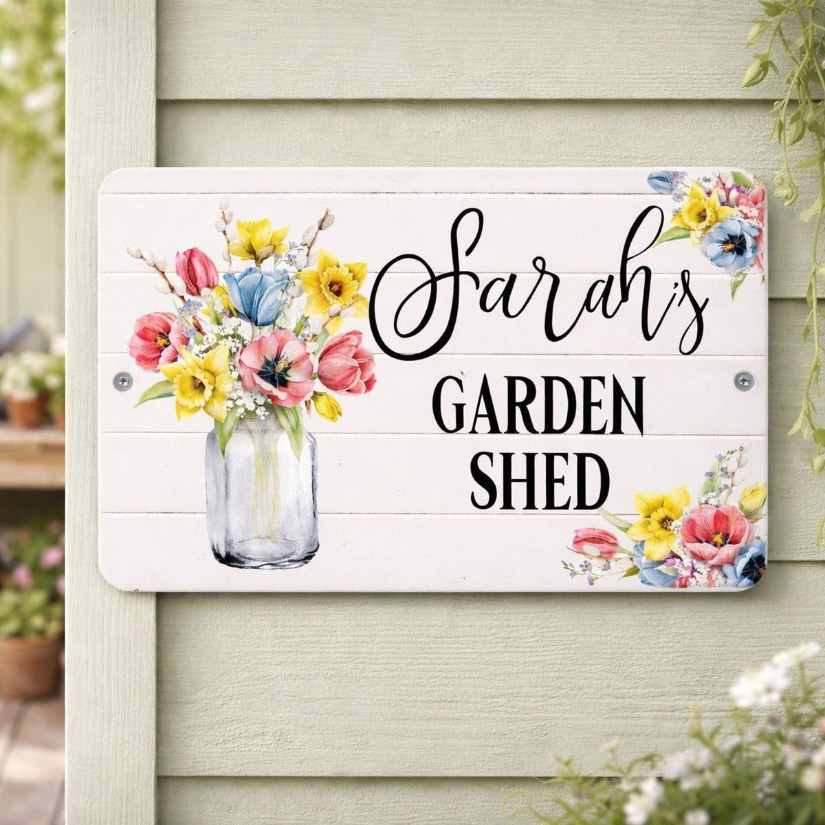A white sign with a vase of colorful flowers and the text "Sarah's Garden Shed" on it, mounted on a wooden wall.