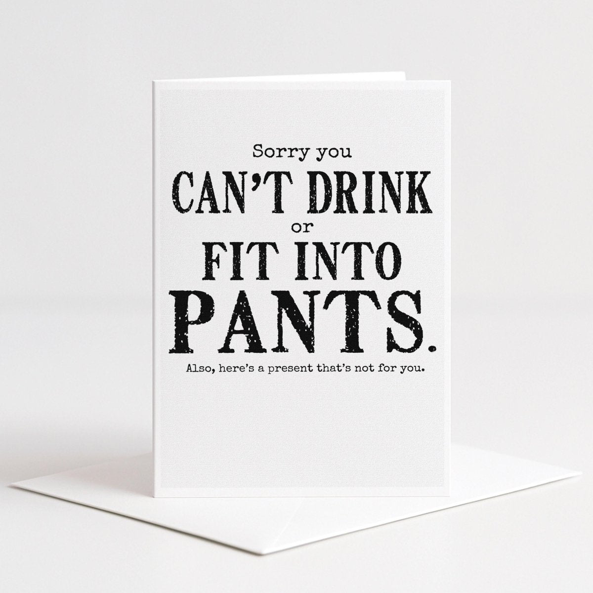 White 5-by-7 greeting card displayed upright on a white envelope with distressed black typography reading, “Sorry you can’t drink or fit into pants. Also, here’s a present that’s not for you.”