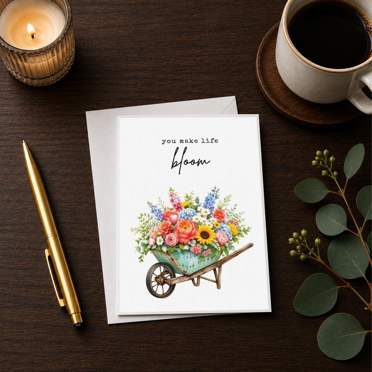 a card with a floral design, a pen, a candle, and a cup of coffee on a wooden surface.