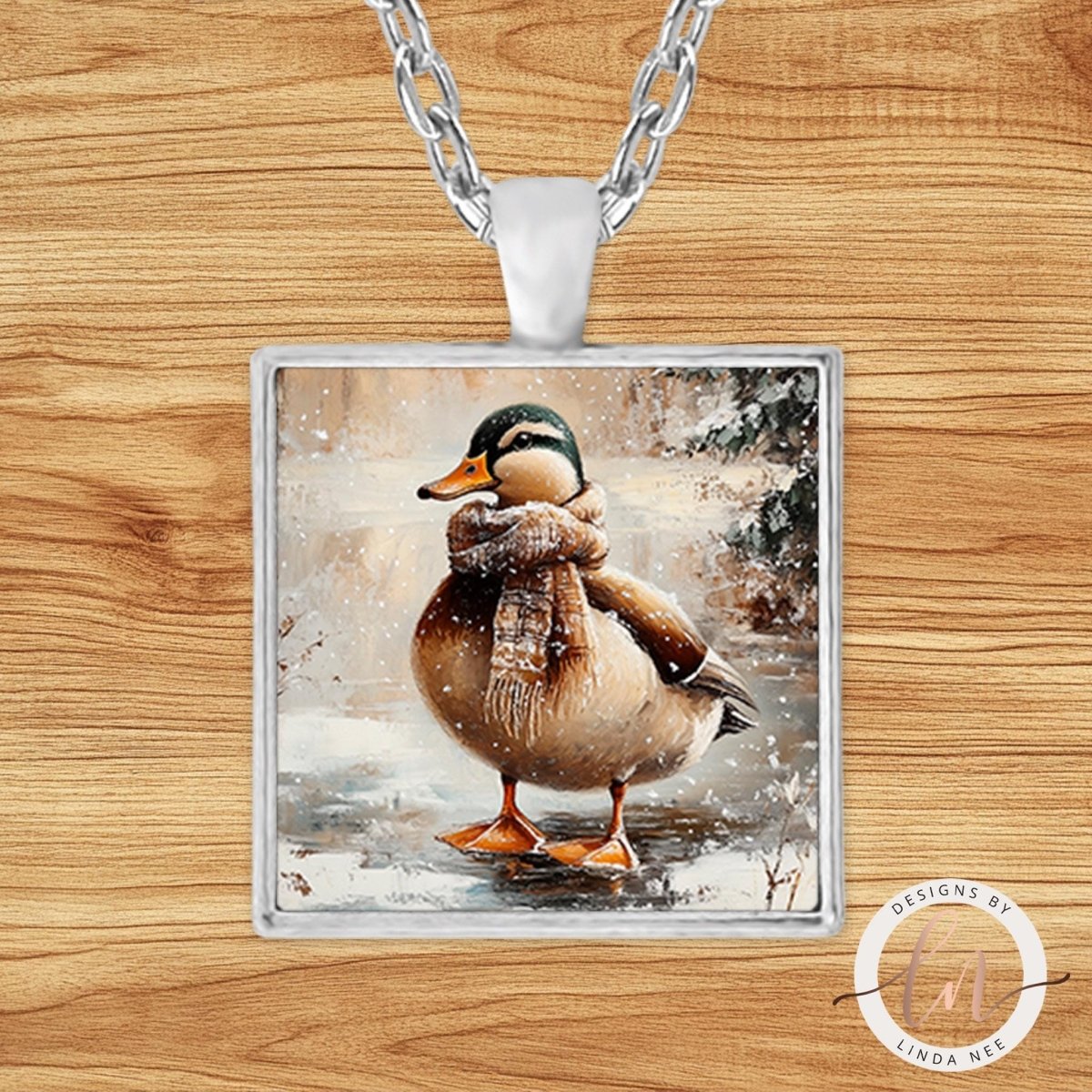 A pendant necklace with a square glass pendant featuring a duck wearing a scarf and standing in a snowy landscape.