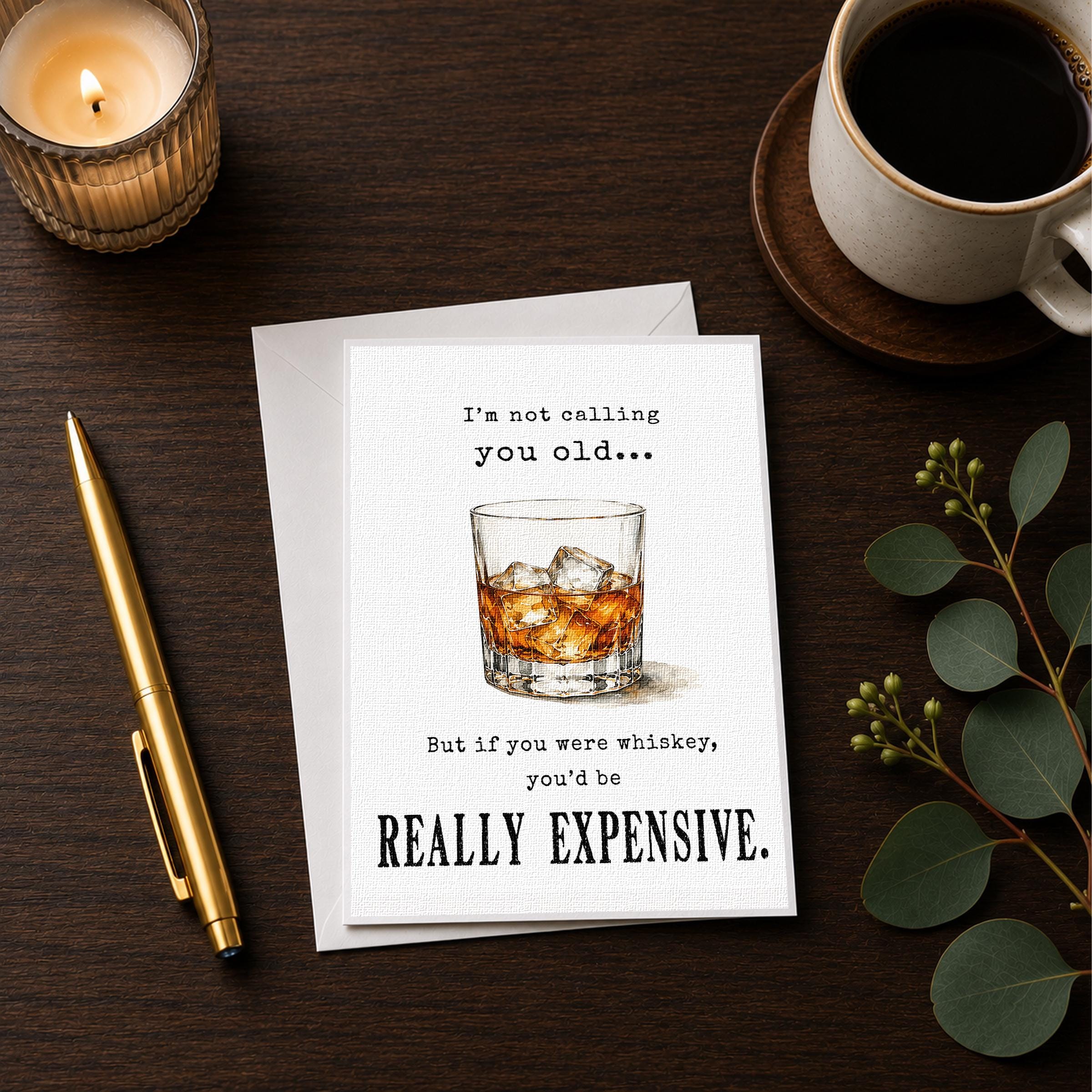 a card with a humorous message, a glass of whiskey, a candle, a pen, and a cup of coffee on a wooden surface.