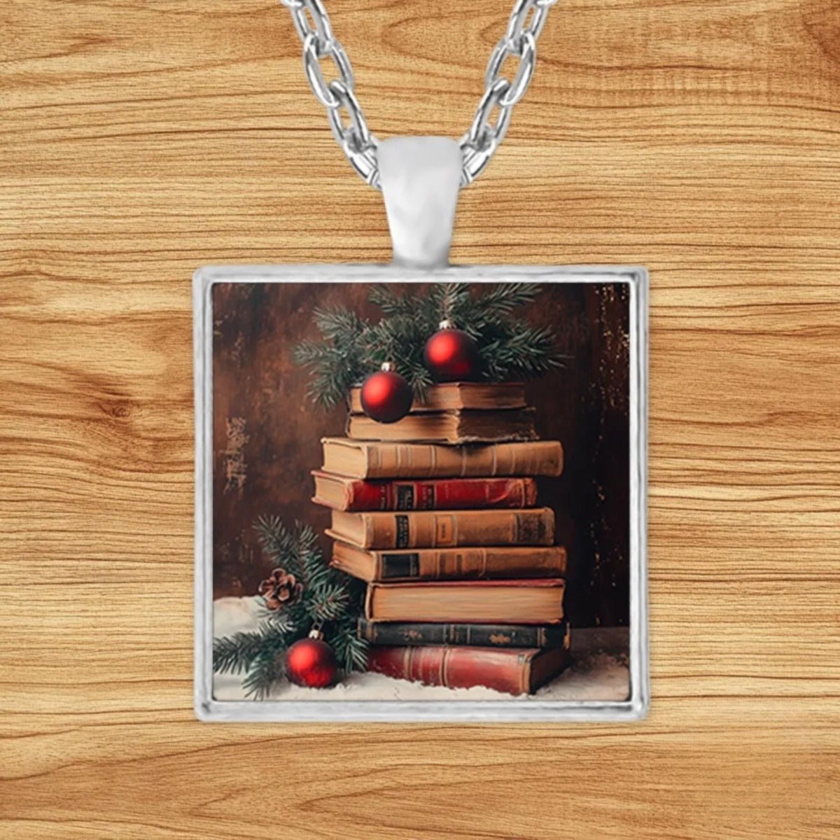 Square silver charm pendant necklace featuring a stack of antique books topped with evergreen sprigs, red ornaments, and snow