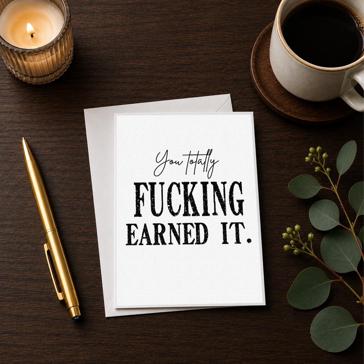 a card with the text "You totally FUCKING EARNED IT." along with a pen and a candle on a wooden surface.