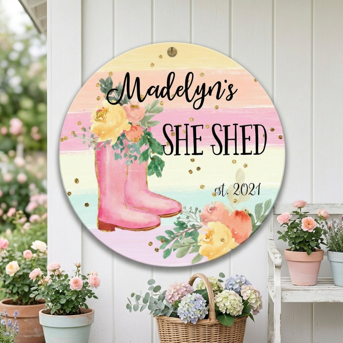 A round sign with a colorful floral design and the text "Madelyn's SHE SHED st. 2021" is displayed on a white wall, surrounded by potted plants and flowers.