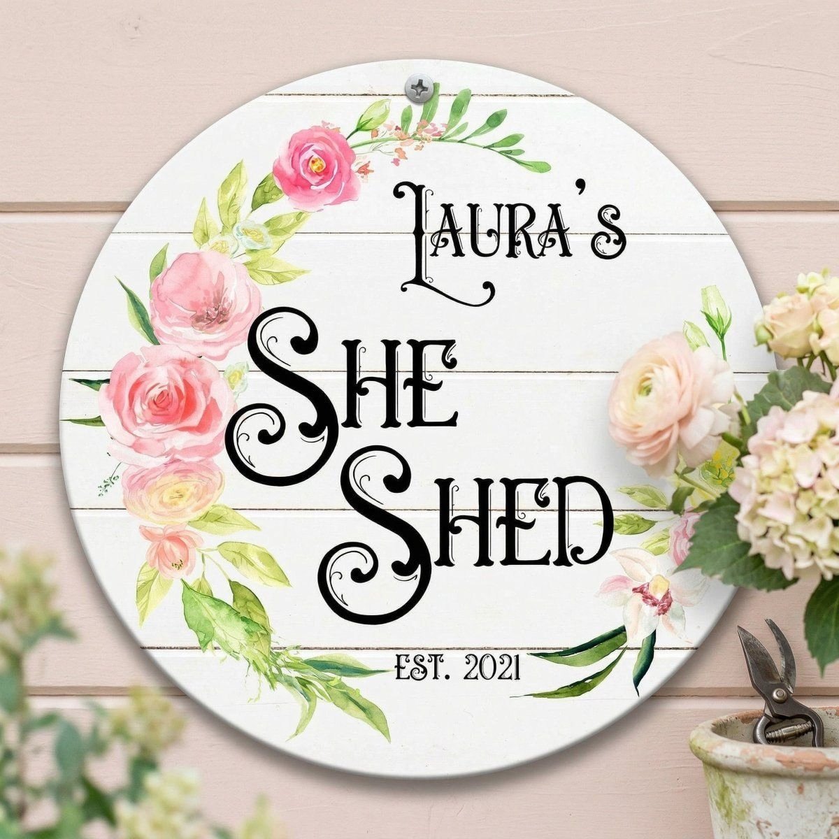A round metal sign with a floral design and the text "Laura's She Shed" displayed on it.