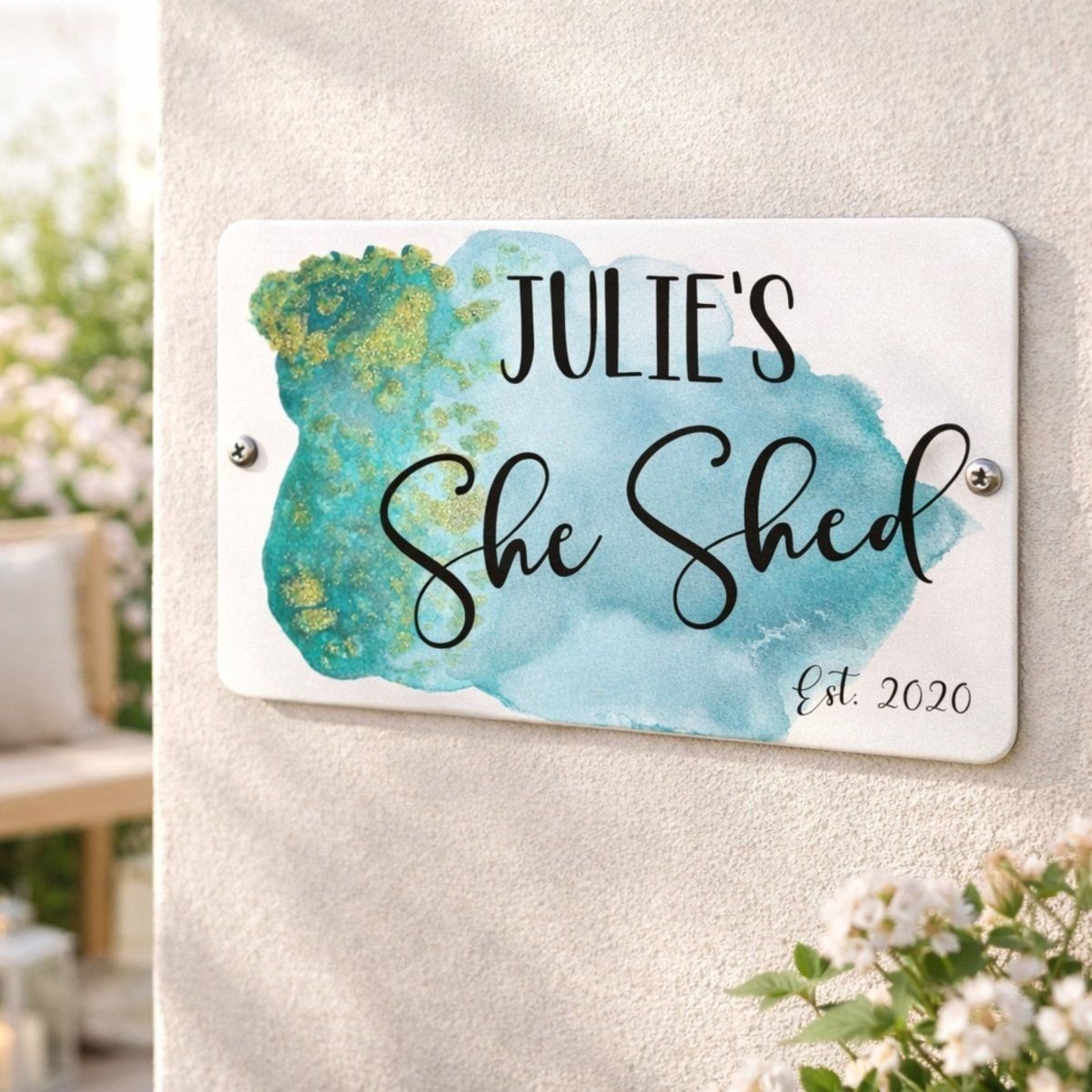 A white sign with blue and green watercolor-like paint, displaying the text "JULIE'S She Shed" and "Est. 2020".
